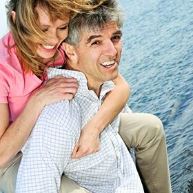 a woman in a pink shirt on the back of a man in a white shirt standing on a beach with the ocean behind them