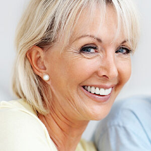 a woman in profile smiling and looking sideways at the camera, with blonde hair and pearl earrings