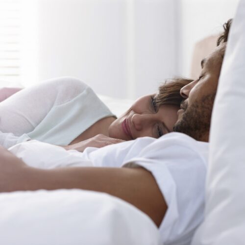 a woman and a man both wearing white lying cuddling in a bed made up of white sheets and pillows