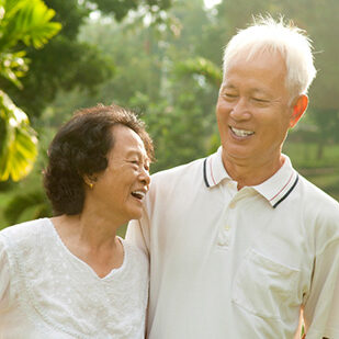 an elderly Asian couple looking at each other and smiling with trees in the background