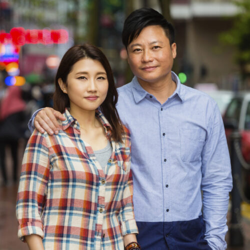 a woman in a plaid shirt standing beside a man in a blue button down shirt, he has his arm around her