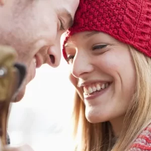 a woman in a red beanie looking at a man and both are smiling at each other, her blonde hair is sticking out from beneath the beanie